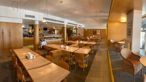 Un restaurant avec des tables et des chaises en bois, un espace cuisine à l’arrière-plan et une ligne jaune délimitant la zone où sont placées les tables.