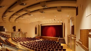 Une salle de conférence avec des rangées de chaises, une scène avec un rideau rouge et un escalier menant à l’étage.