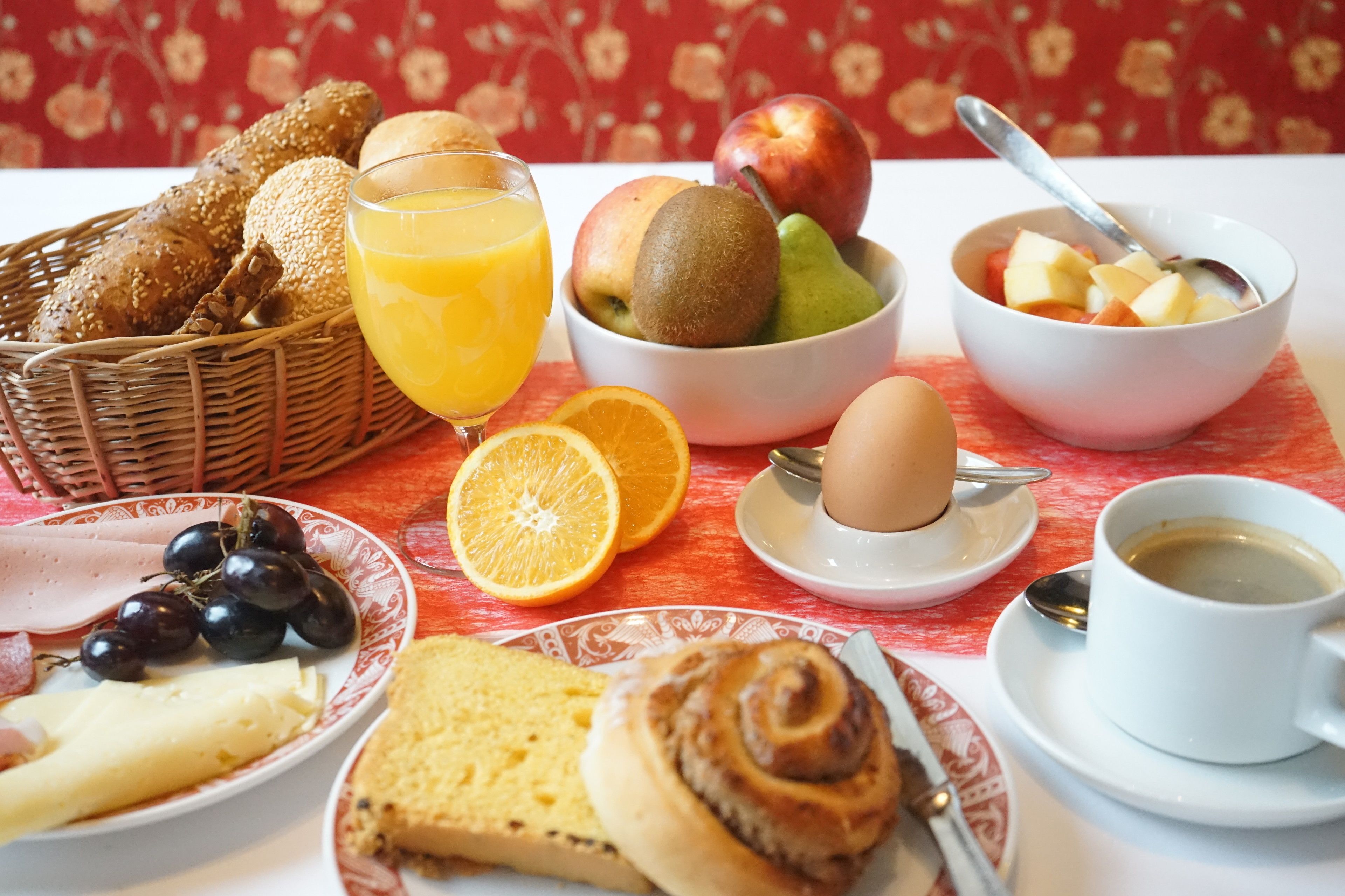 A breakfast table with bread, fruit, a glass of orange juice, a bowl of cereal, a plate of ham and cheese, and a cup of coffee.