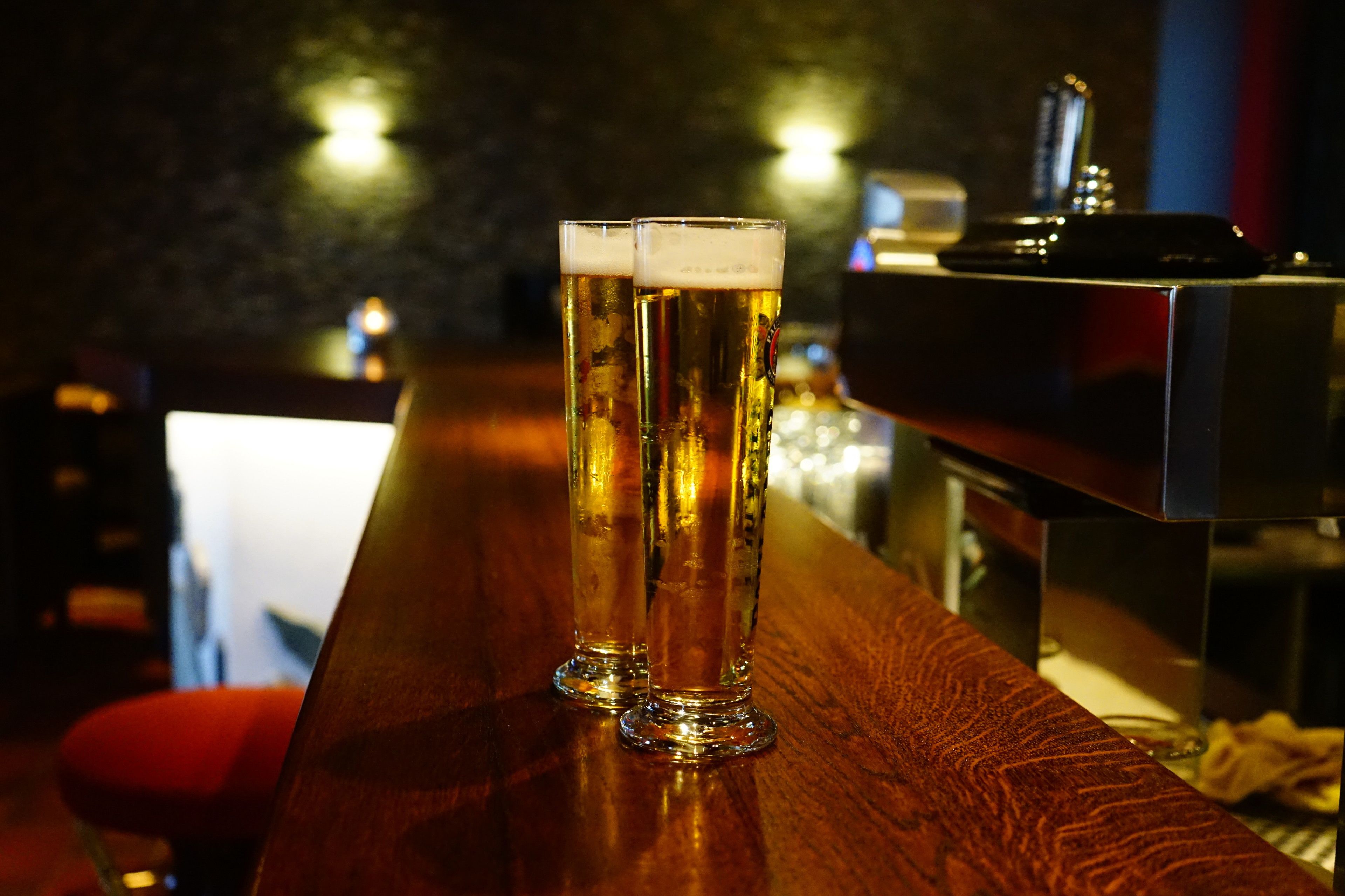 Two tall glasses of beer on a wooden bar counter with a dark background and a barista machine.