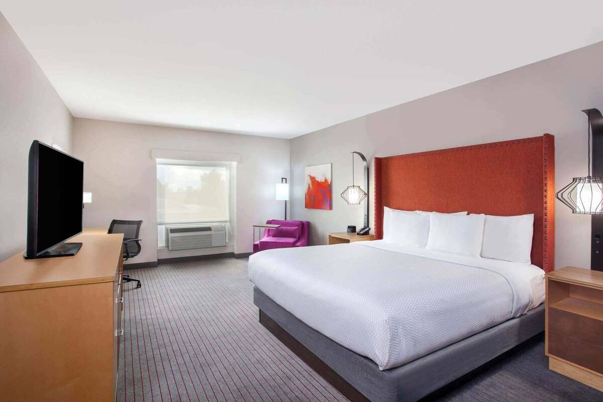 A hotel room with a large bed, a desk with a TV, a chair, a nightstand, and a window.