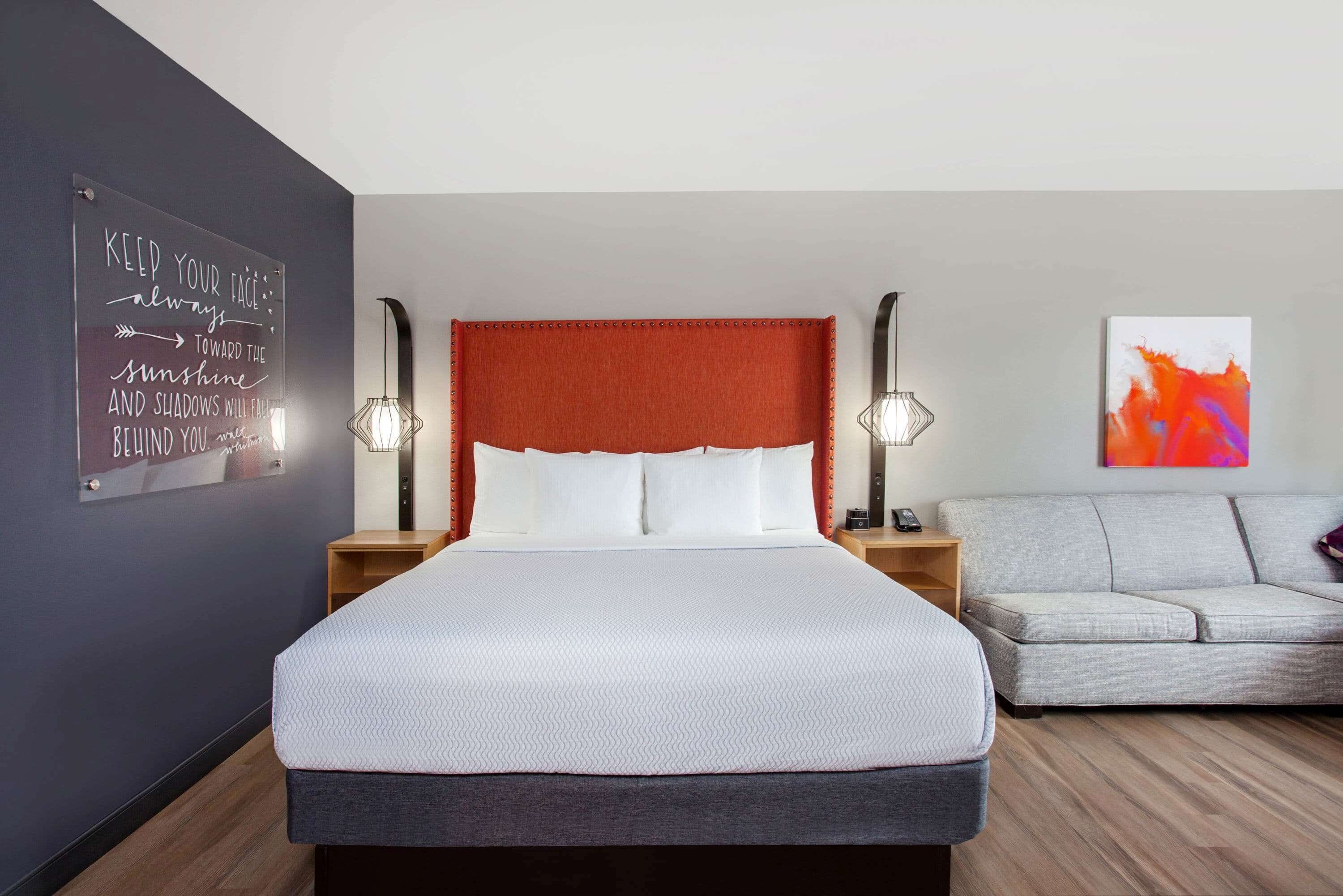 A modern hotel room with a large bed, a grey sofa, a wall-mounted artwork, and a motivational quote on the wall.