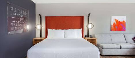 A modern hotel room with a large bed, a grey sofa, a wall-mounted artwork, and a motivational quote on the wall.