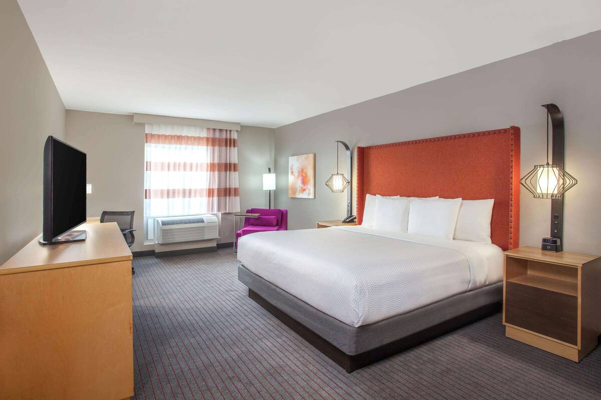 A hotel room with a large bed, a desk with a flat-screen TV, a chair, a bedside table with a lamp, and a window with curtains.