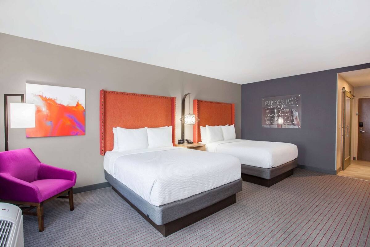 A hotel room with two beds, a chair, a wall art piece, and a motivational quote on the wall.
