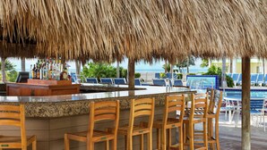 Un área de bar con techo de paja, sillas de madera y vista a la piscina.