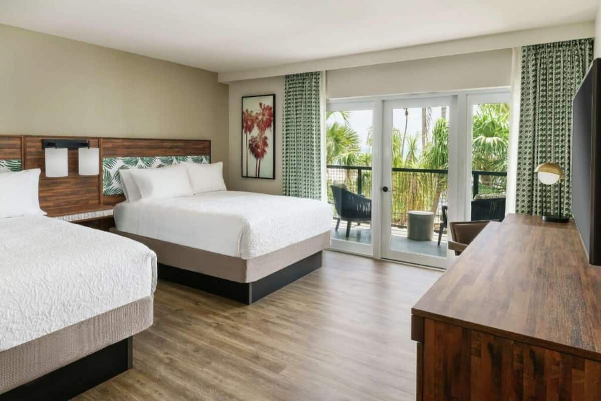 A hotel room with two beds, a wooden desk, a balcony with a chair, and a view of palm trees.