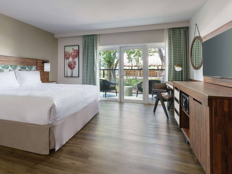 Habitación de hotel con una cama grande, un escritorio con silla, televisión y balcón con sillas.
