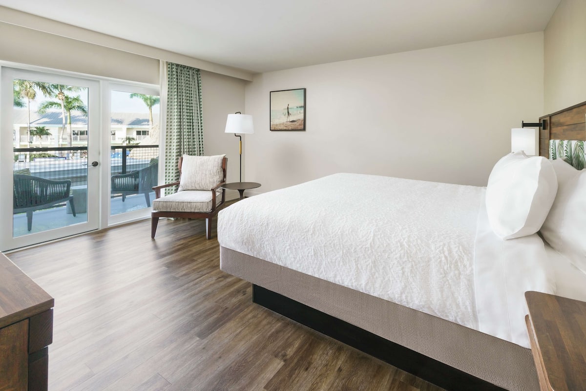 A hotel room with a large bed, a chair, a desk, and a view of a pool and palm trees through a sliding door.