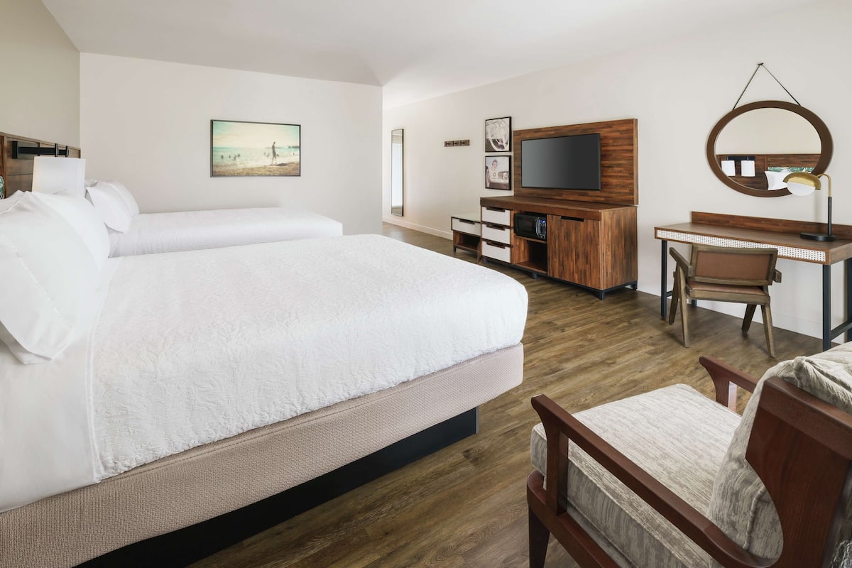 A hotel room with two beds, a desk, a chair, a TV, and a large mirror.