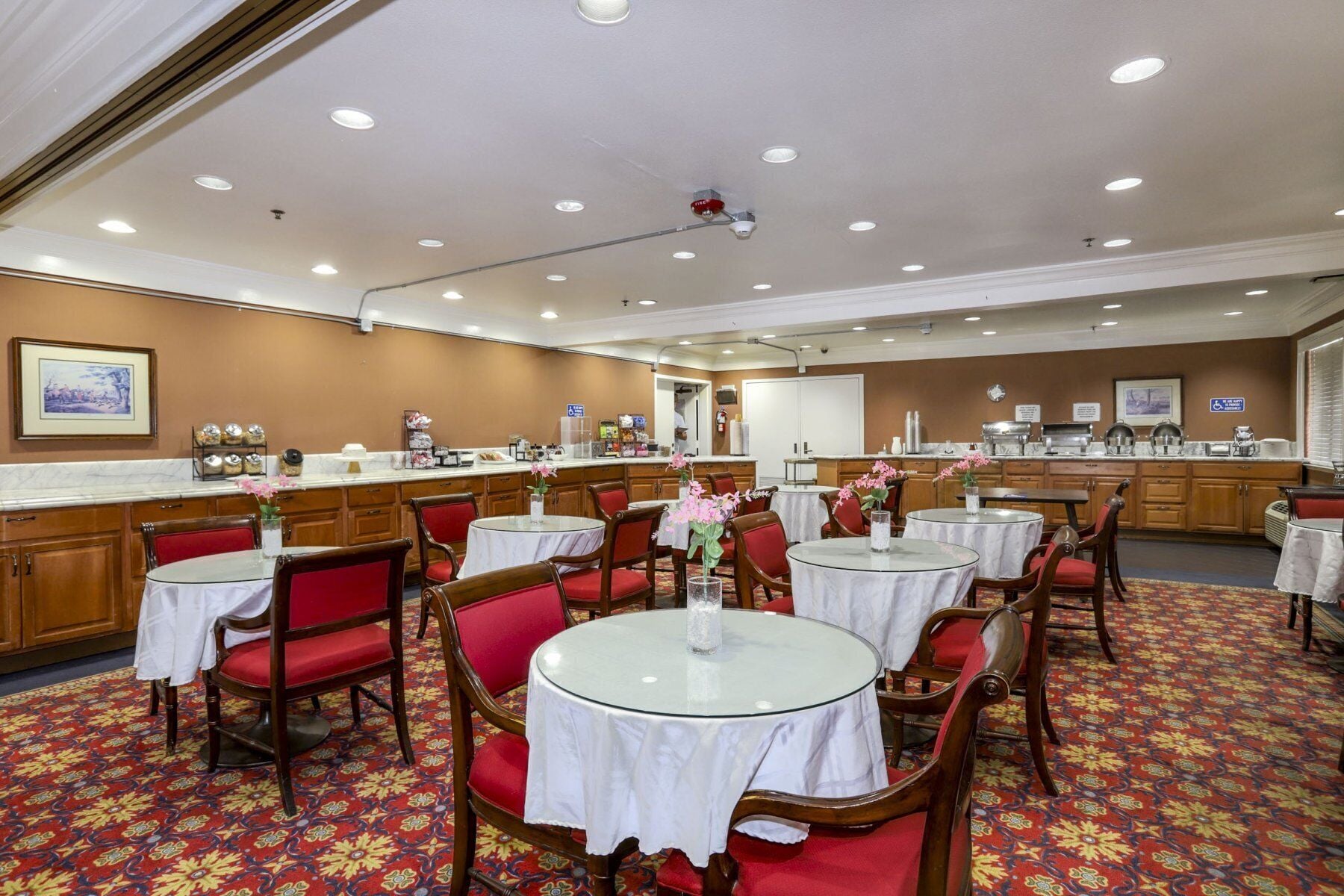 A hotel dining area with round tables, red chairs, a buffet counter, and a patterned carpet.