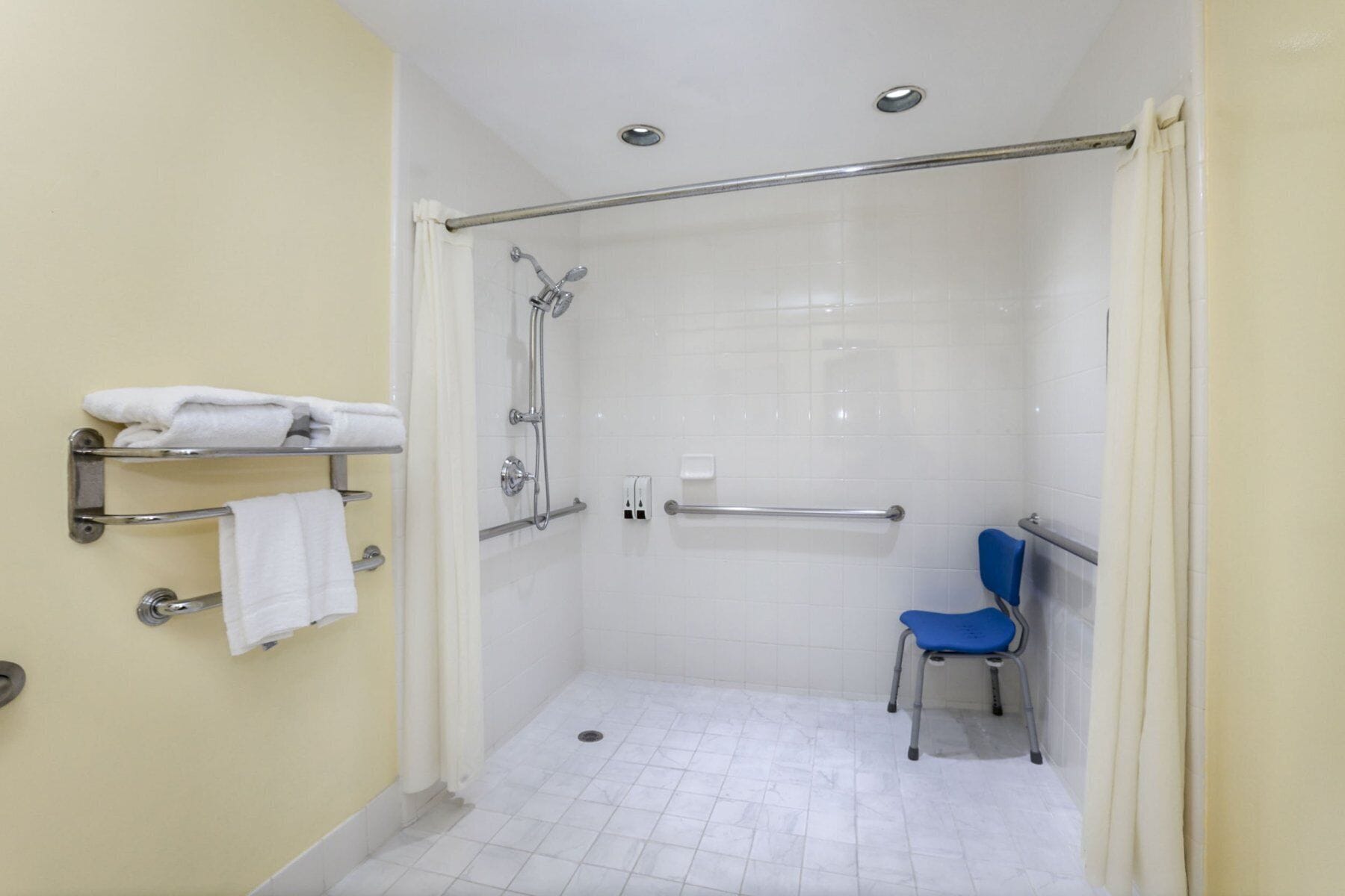 Un petit espace de douche fermé, équipé d’une barre d’appui, d’une chaise bleue et d’un porte-serviettes avec des serviettes.