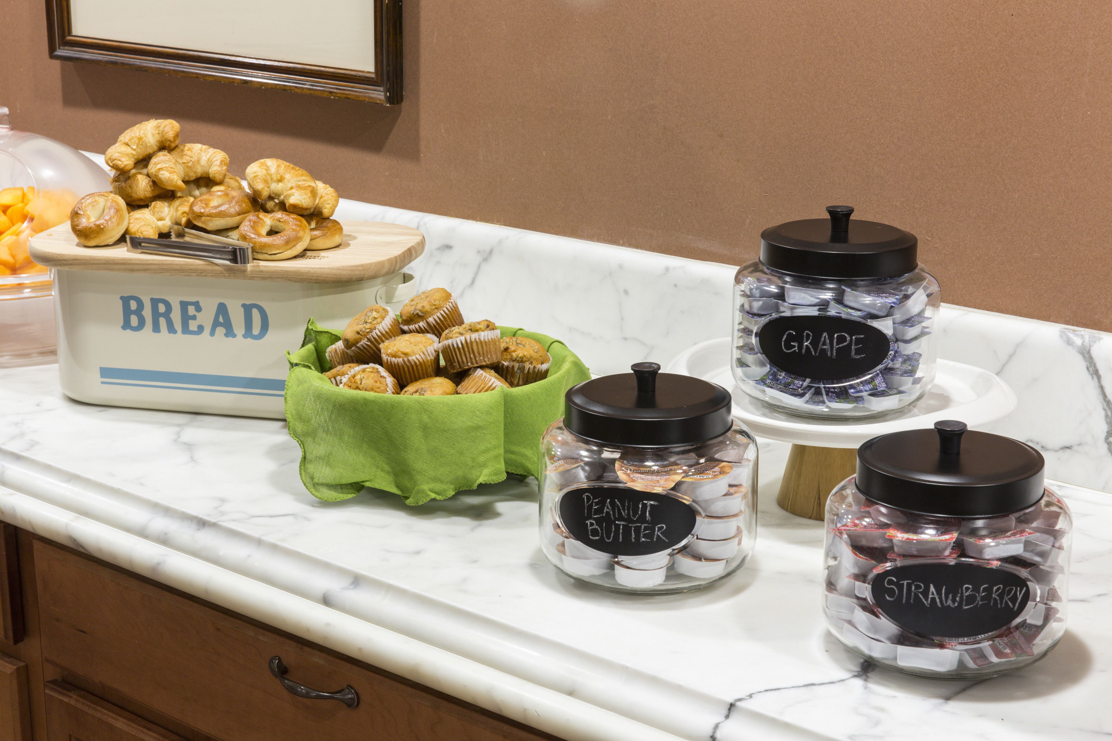 A kitchen counter with labeled jars of peanut butter, grape, and strawberry preserves, a bowl of croissants, and a bread container.