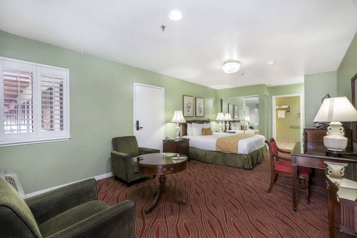 A hotel room with a bed, two chairs, a desk, a mirror, and a window with blinds.