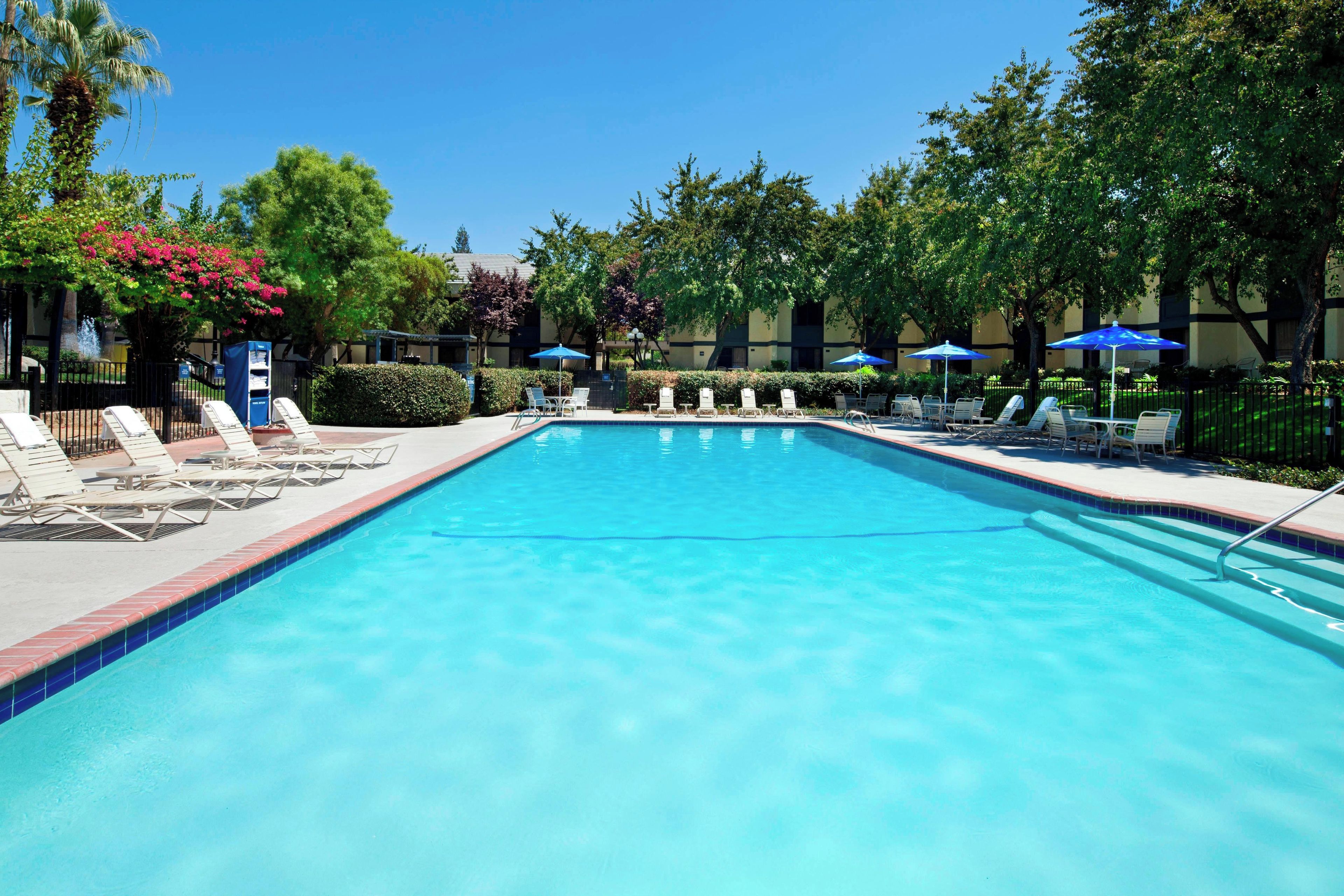 A swimming pool with lounge chairs and umbrellas, surrounded by trees and a building.