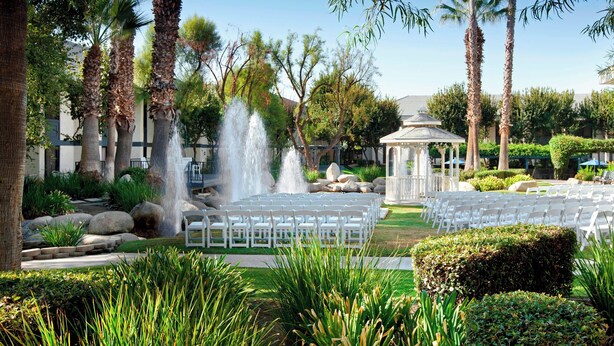An outdoor wedding venue with a fountain, palm trees, and white chairs arranged for guests. - Four Points by Sheraton Bakersfield (Bakersfield)