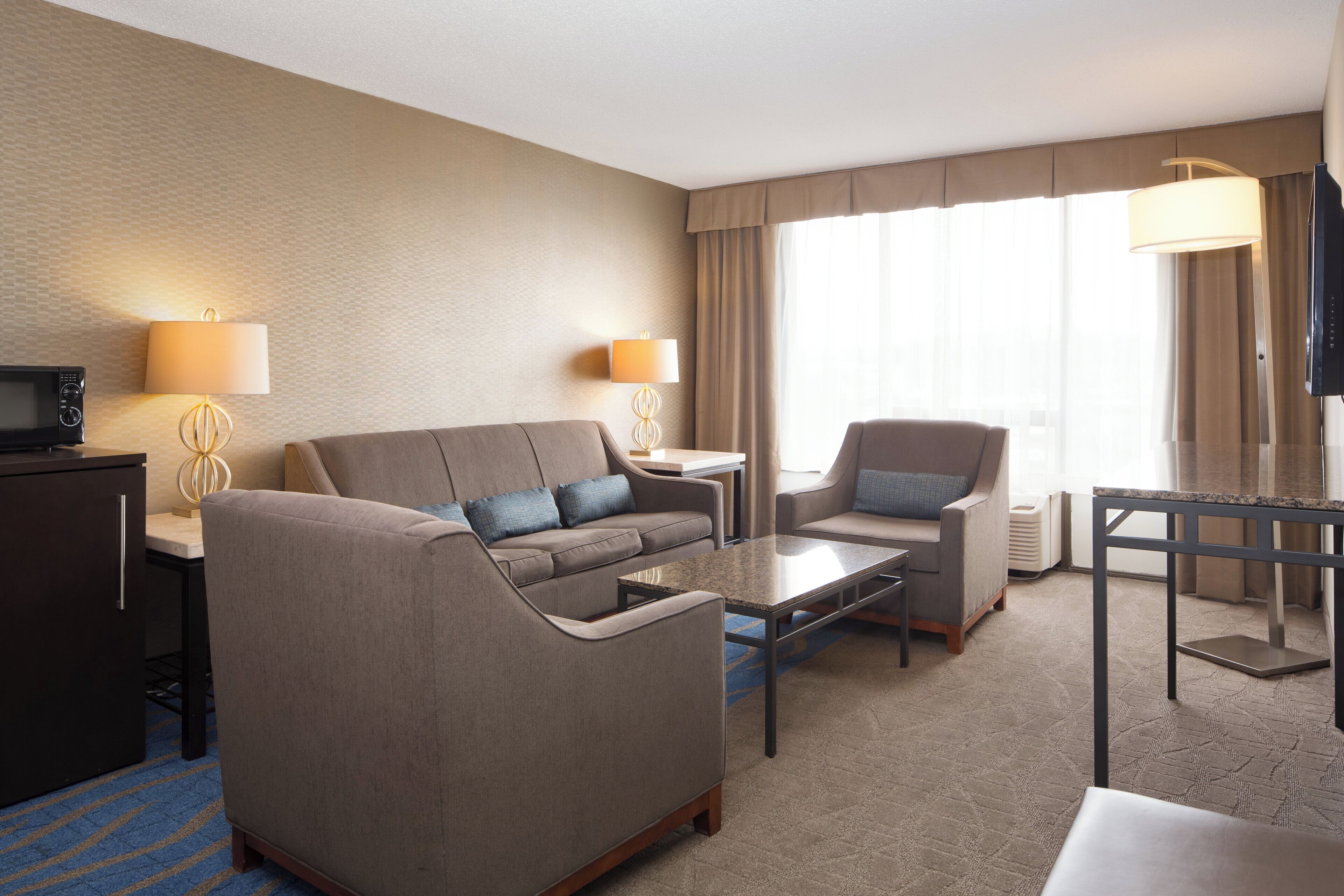 A hotel room with a sofa, armchair, coffee table, TV, and lamps.