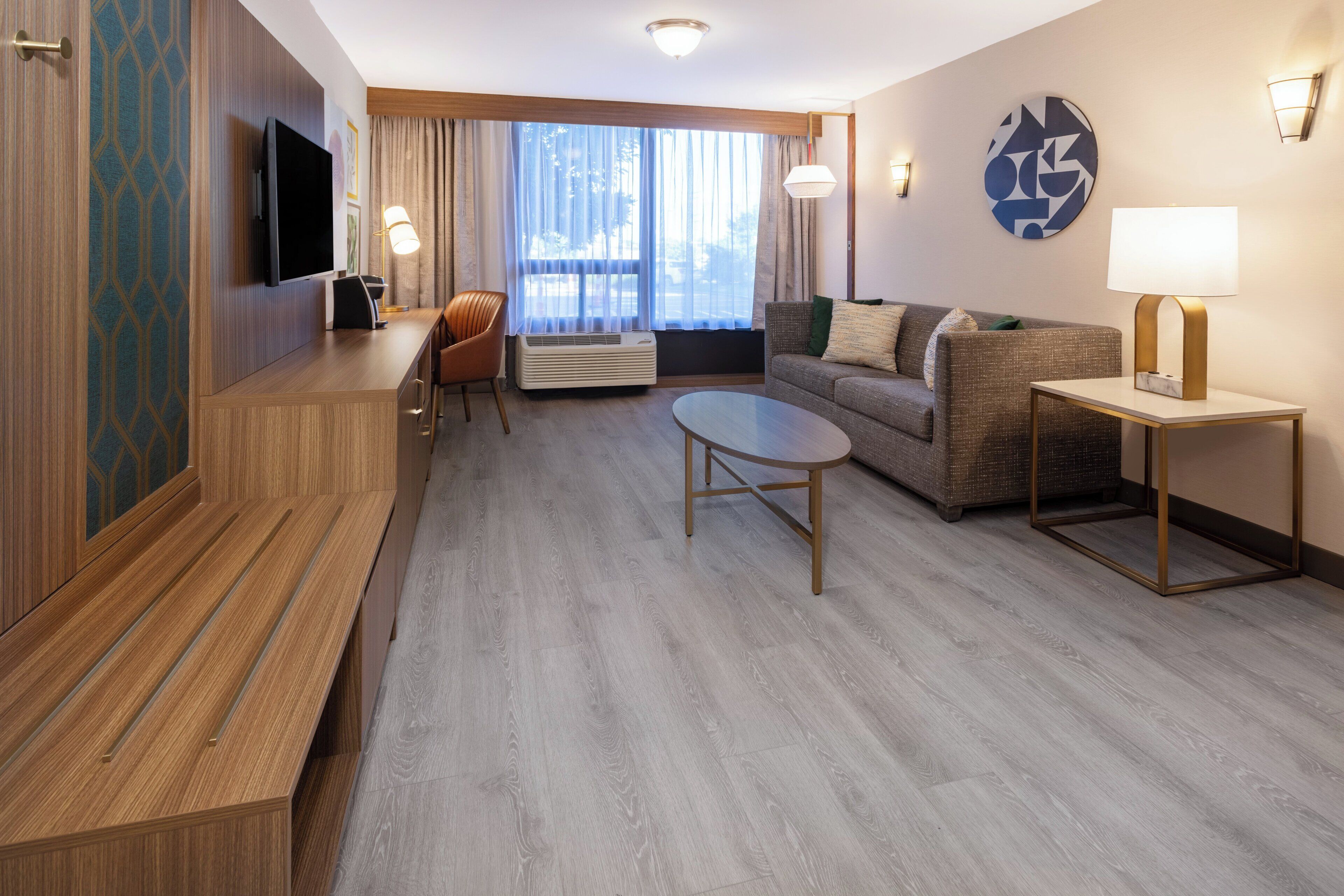 A hotel room with a flat-screen TV, a sofa, a coffee table, a chair, and a desk with a lamp.