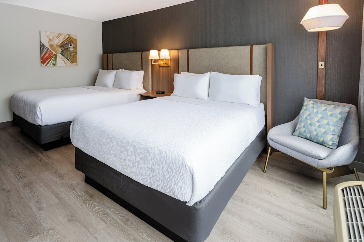 A hotel room with two beds, a chair, and a wall-mounted lamp.