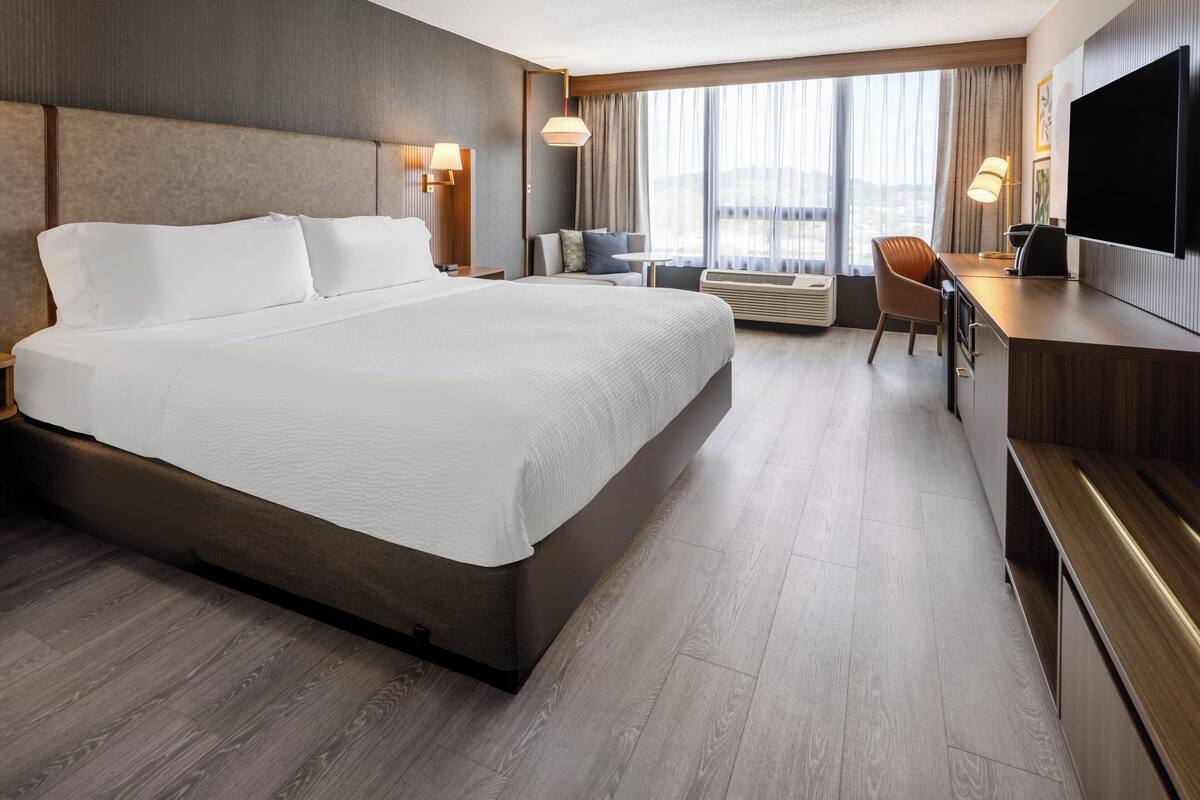A hotel room with a large bed, a desk, a chair, a TV, and a window with curtains.