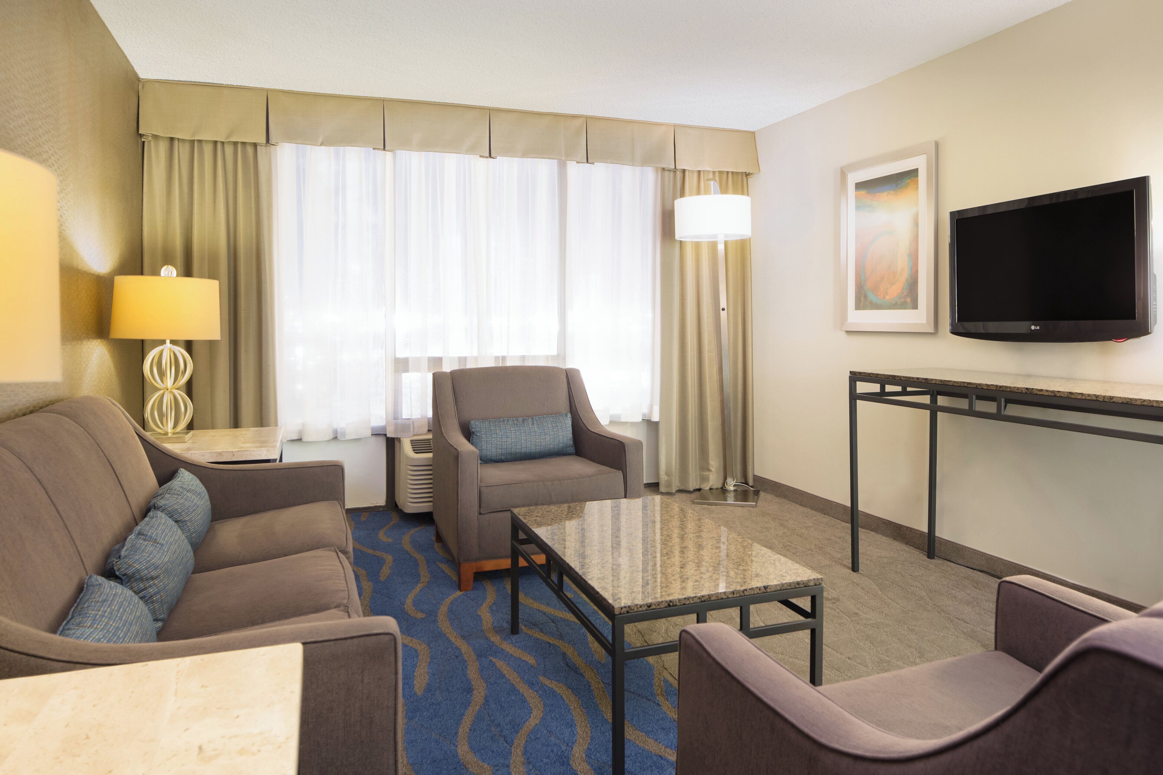 A hotel room with a sofa, armchair, a small table, a TV, and a lamp.