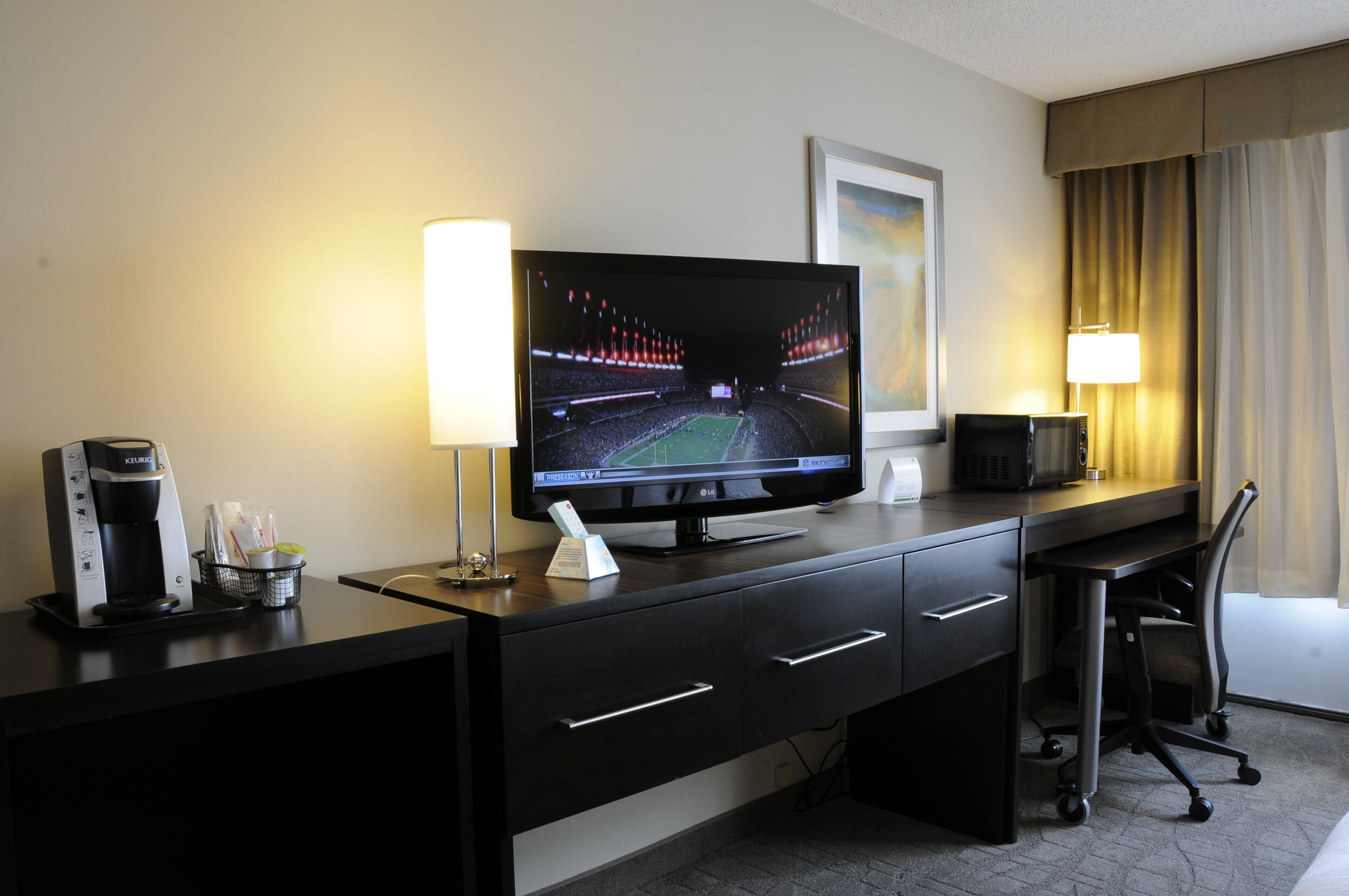 Habitación de hotel con televisión de pantalla plana, cafetera, control remoto, escritorio con cajones, silla y lámpara.