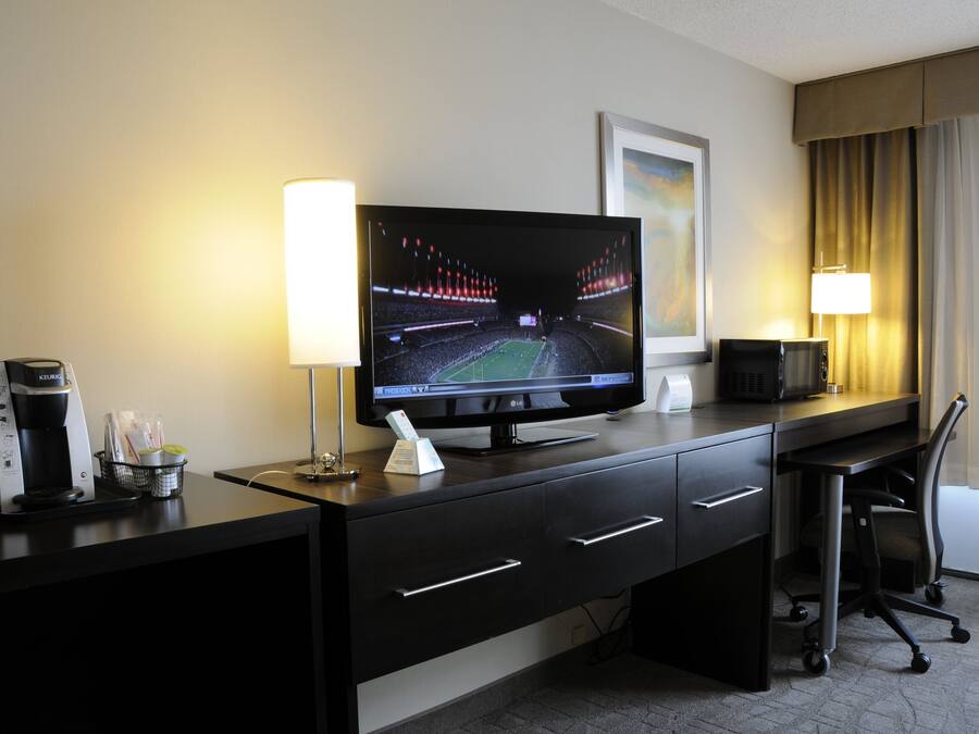 Habitación de hotel con televisión de pantalla plana, cafetera, control remoto, escritorio con cajones, silla y lámpara.