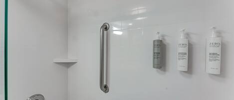 A shower area with a handheld showerhead, a grab bar, and three bottles of ZERO% shampoo and conditioner.
