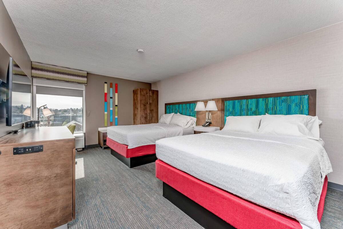 A hotel room with two beds, a desk, a window, and a view of the city.