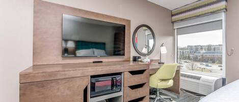 A hotel room with a wooden desk, a flat-screen TV, a mirror, a chair, a bed, and a large window with a city view.