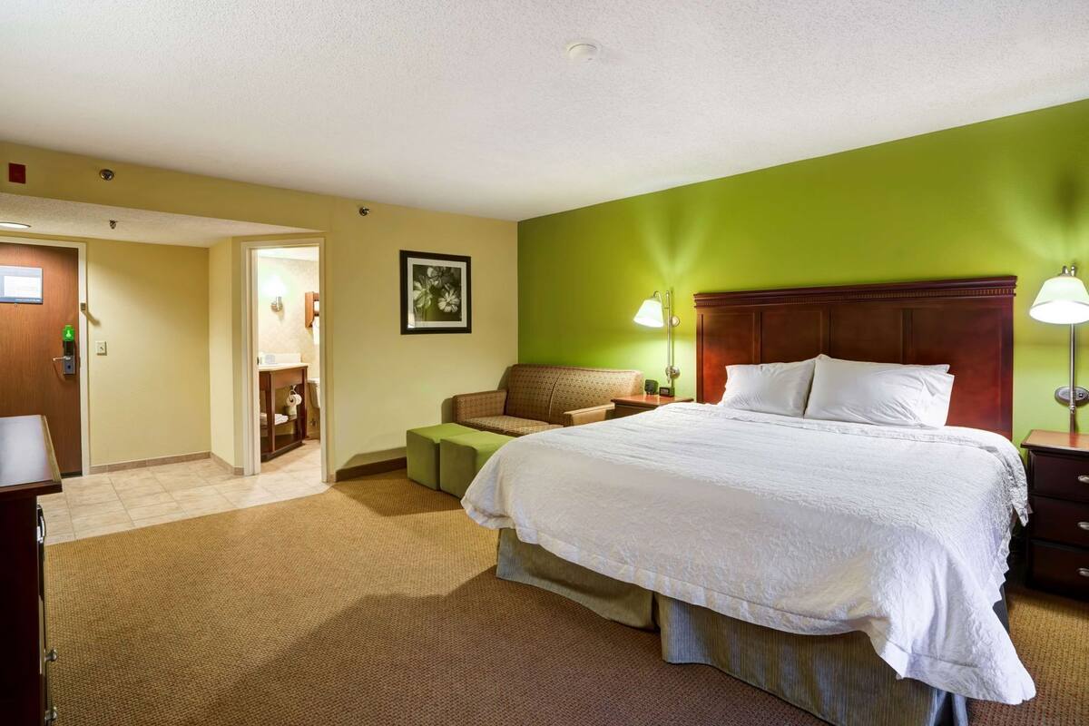 A hotel room with a large bed, a green accent wall, a wooden headboard, a bedside table with a lamp, and a small seating area with a couch and ottoman.