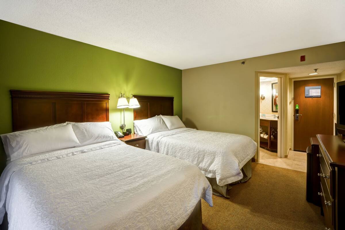 A hotel room with two beds, a nightstand with a lamp, a dresser, and a bathroom visible through an open door.