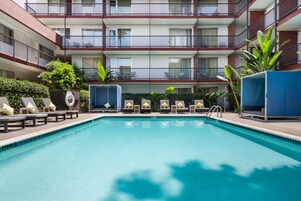 Una piscina con tumbonas y un edificio de varios pisos con balcones.