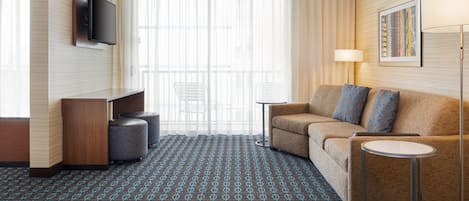 A hotel room with a sofa, a TV mounted on the wall, a small table, a lamp, and a patterned carpet.