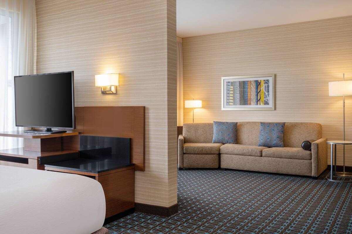 A hotel room with a flat-screen TV, a sofa with patterned cushions, a framed artwork on the wall, and a patterned carpet.
