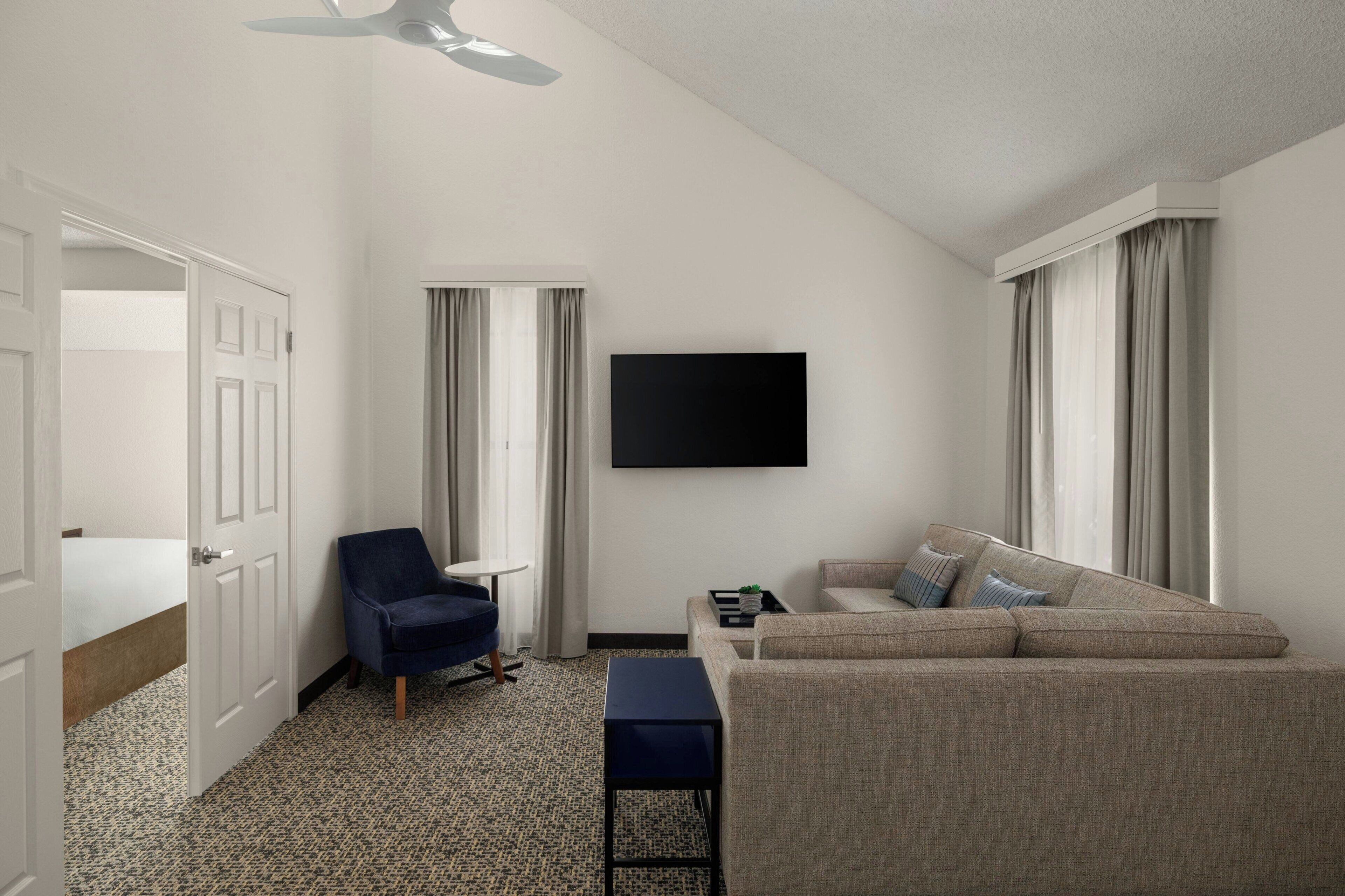 A hotel room with a bed, a blue armchair, a beige sofa, a small table, and a flat-screen TV mounted on the wall.