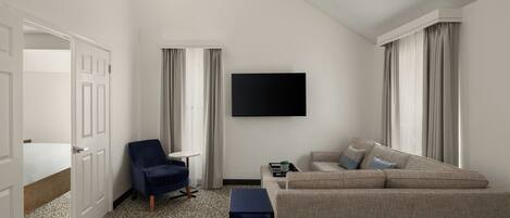 A hotel room with a bed, a blue armchair, a beige sofa, a small table, and a flat-screen TV mounted on the wall.