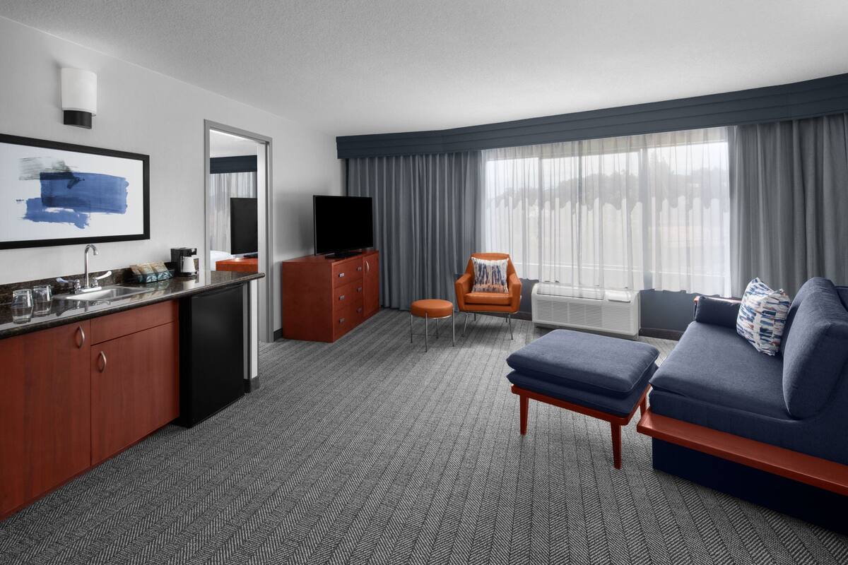 A hotel room with a kitchenette, a sofa, a chair, and a large window with curtains.