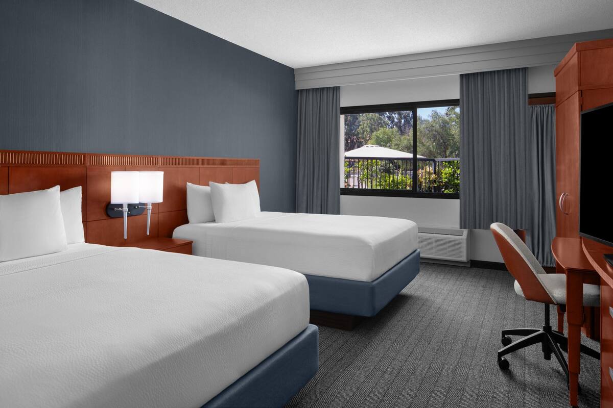 A hotel room with two beds, a desk, a TV, and a window with a view of greenery.