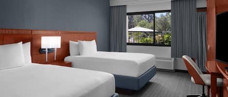 A hotel room with two beds, a desk, a TV, and a window with a view of greenery.
