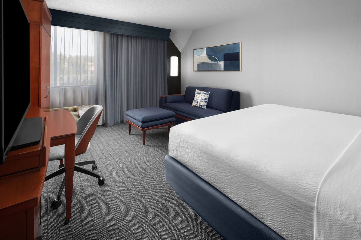 A hotel room with a bed, a desk with a chair, a sofa, and a window with curtains.