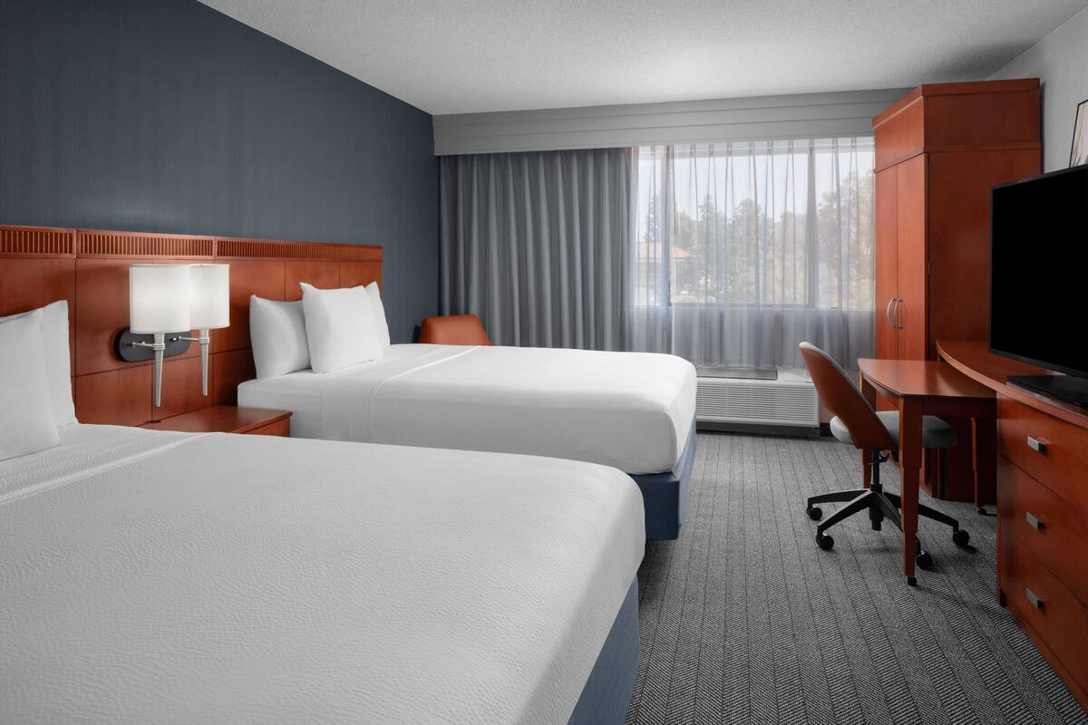 A hotel room with two beds, a desk, a television, and a large window with curtains.