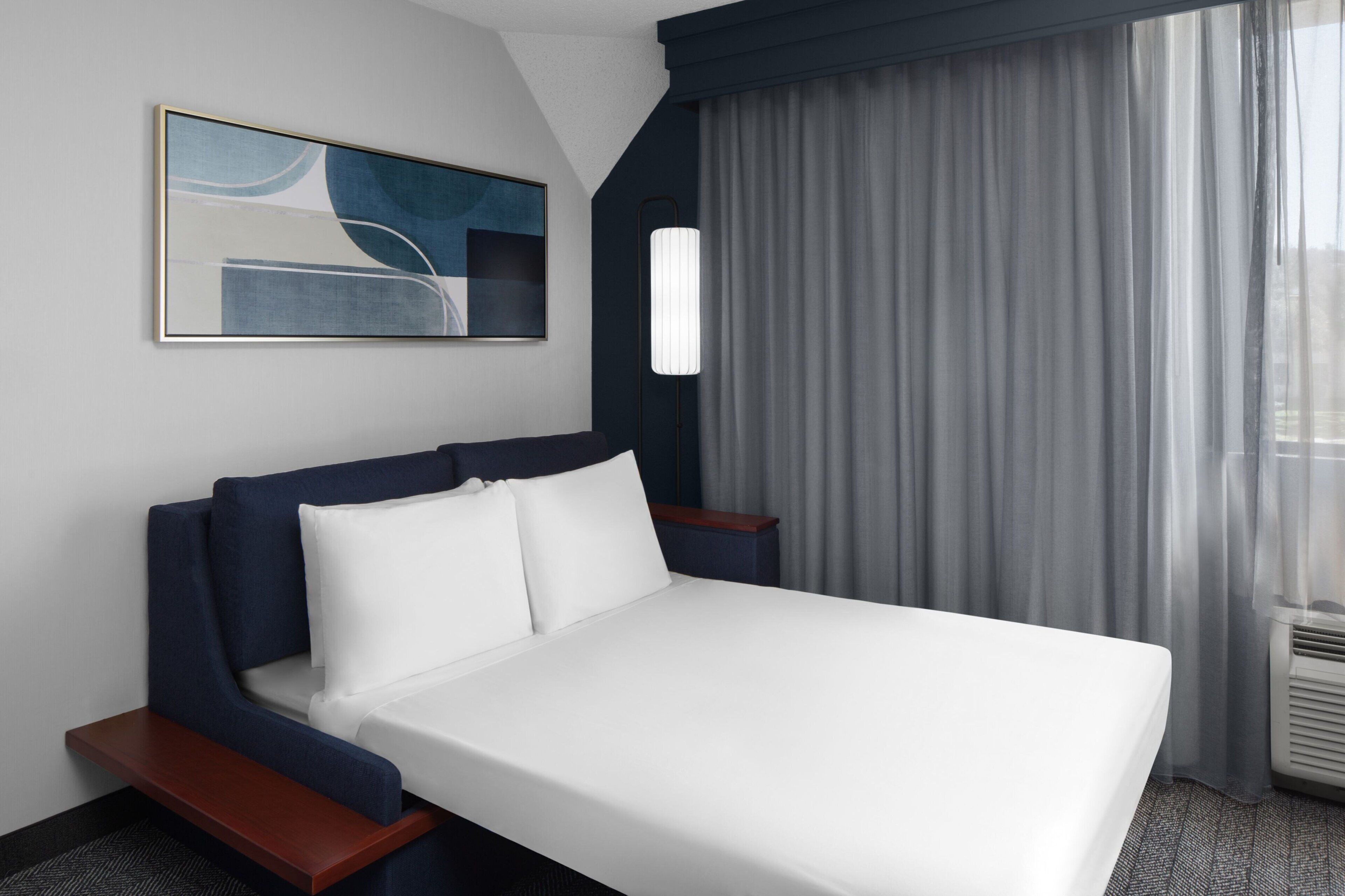 A hotel room with a bed, a bedside table, a lamp, a painting on the wall, and a window with curtains.