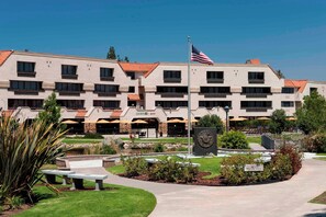 Hotel features a modern design with a flat roof and windows on each floor, surrounded by a landscaped area with a seating arrangement and a flagpole.