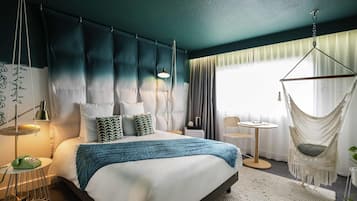 Ein modernes Hotelzimmer mit einem großen Bett, einem Hängekorbstuhl, einem kleinen Tisch und Blick ins Freie.