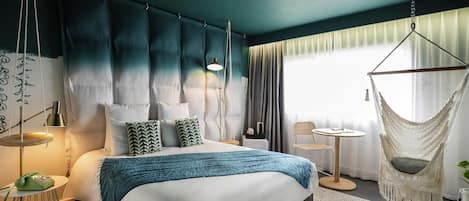 A modern hotel room with a large bed, a hanging chair, a small table, and a view of the outdoors.