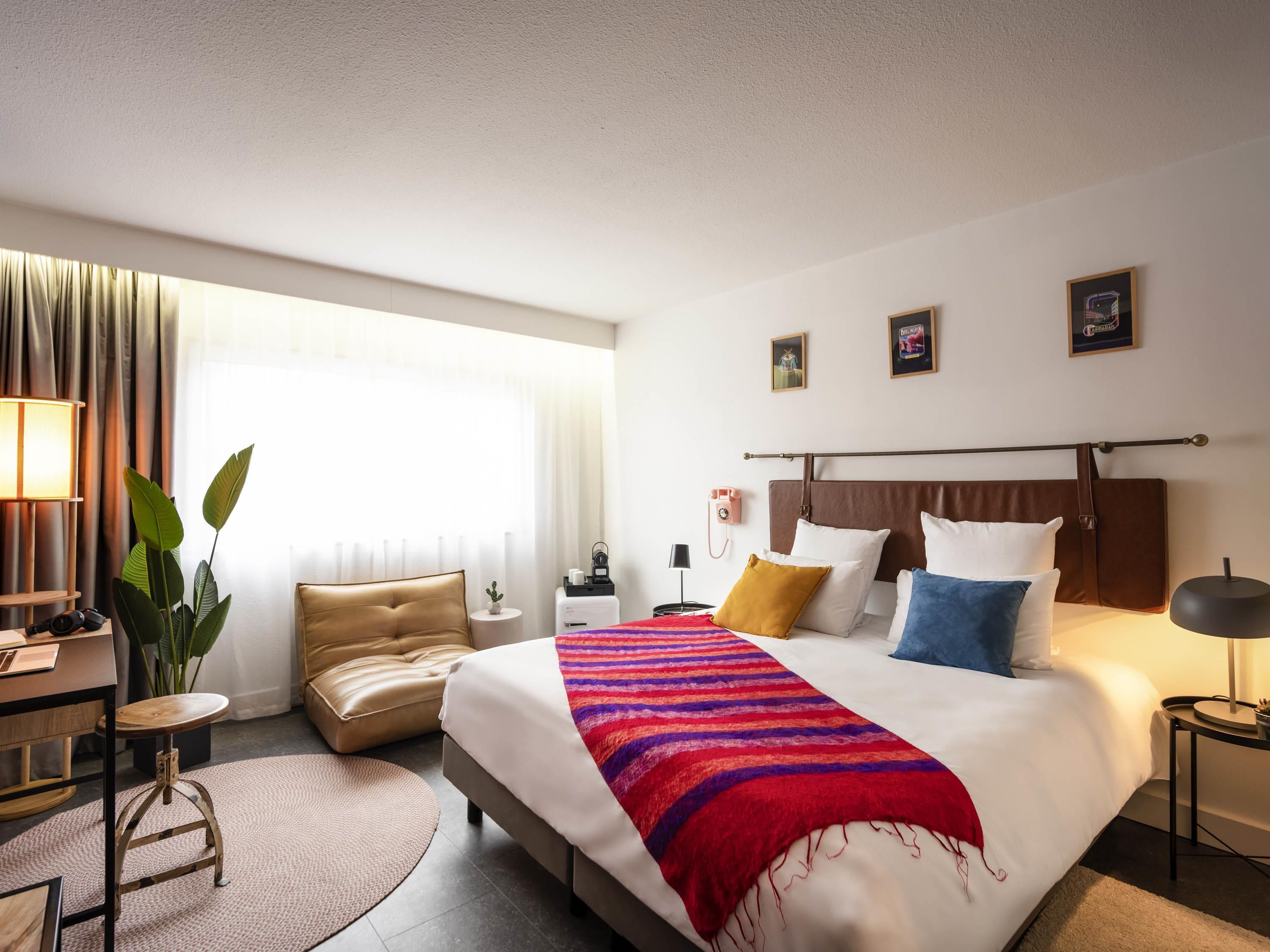 Habitación de hotel con una cama grande, un escritorio, una silla, una planta en maceta y una ventana con cortinas.