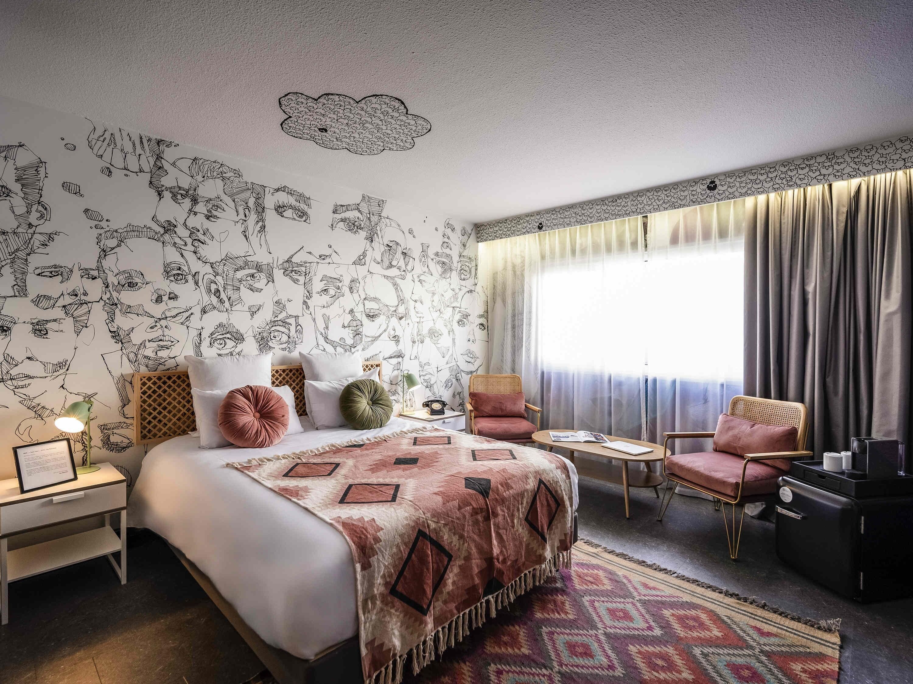 A hotel room with a bed, two chairs, a small table, a nightstand, and a window with curtains.