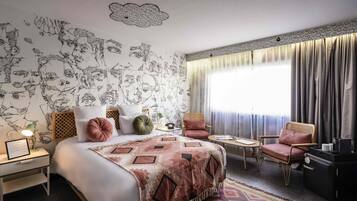 A hotel room with a bed, two chairs, a small table, a nightstand, and a window with curtains.