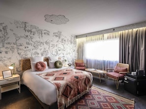 Habitación de hotel con cama, dos sillas, una mesita, una mesita de noche y una ventana con cortinas.
